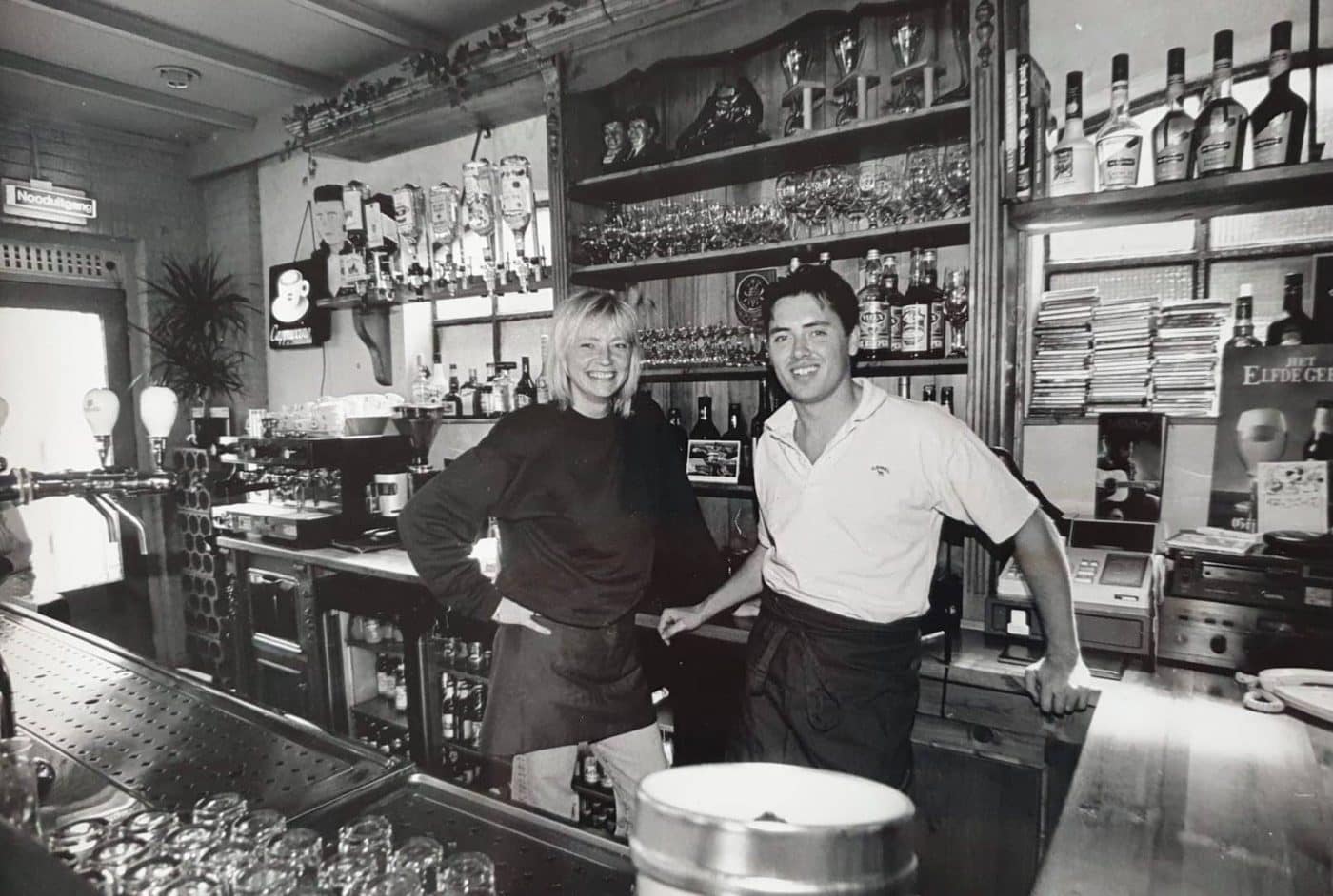 De eigenaren lachend achter de bar van Café Roosje, zwart-witfoto